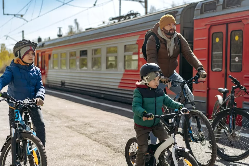 Man and two children on bikes next to red and silver train on sunny day.